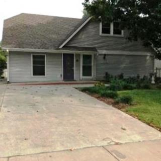 A grayish multi-story residence with a double window on upper level and two single windows on ground level and large concrete driveway.